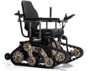 All-terrain wheelchair with tank-like tracks, camouflage coloring, padded seat, armrests, joystick controller, and sturdy frame designed for outdoor mobility over rough surfaces.
