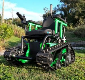 A green and black all-terrain wheelchair with tank-like tracks is parked on grassy ground outdoors, surrounded by trees and sunlight.