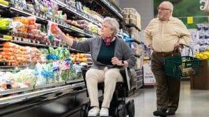 An older woman in a wheelchair selects produce from a grocery store shelf while an older man stands nearby holding a shopping basket. Both are focused on shopping in the fresh produce aisle.