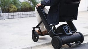 A person in business attire uses a modern, compact electric wheelchair to navigate over a curb on a paved outdoor path, with greenery and a stone wall in the background.