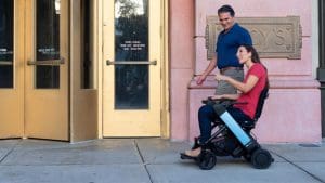 A woman using a motorized wheelchair and a man walking beside her smile and talk as they travel on a sidewalk outside a building with gold doors and a plaque that says CYCYS.