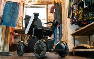 A modern electric wheelchair with large, rugged wheels is positioned in the aisle of a clothing store, surrounded by hanging garments and rustic wooden shelves.