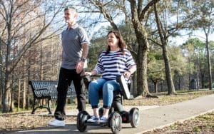 A woman using a motorized wheelchair and a man walking beside her smile as they travel along a paved park path lined with trees and benches on a sunny day.