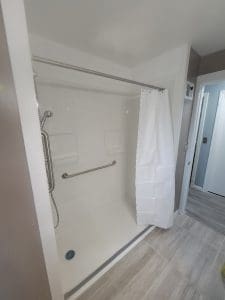 A white walk-in shower with a grab bar, handheld showerhead, and a partially drawn white shower curtain. The bathroom has light gray tiled flooring and beige walls; a doorway leads to a hallway.