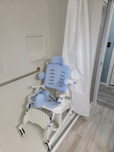 A blue and white shower wheelchair with armrests and padding is positioned inside a walk-in shower with a white curtain, a grab bar, and light-colored tile flooring and walls.