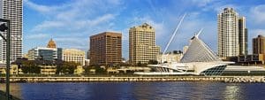 Skyline of Milwaukee, Wisconsin featuring the modern white architecture of the Milwaukee Art Museum on the waterfront, surrounded by tall office buildings under a blue sky.