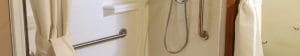A white accessible shower with a handheld showerhead, grab bars, a curtain partially open, and white tiled walls.