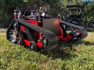 A close-up of a small, red and black tracked robotic vehicle with tank-like treads sitting on grass, surrounded by greenery. Another similar robot is visible in the background.