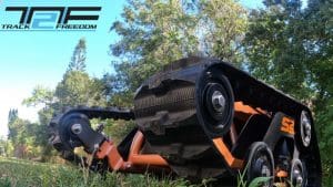 Close-up view of a rugged, all-terrain tracked wheelchair on grass, with the Track 2 Freedom logo in the top left corner. Trees and greenery are visible in the background under a blue sky.
