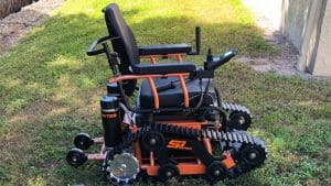 A motorized wheelchair with black and orange accents, equipped with large tank-like rubber tracks and small wheels, designed for off-road or rough terrain, is parked on a grassy area near a building.