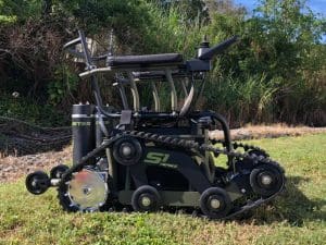 A rugged, all-terrain wheelchair with tank-like tracks designed for outdoor use is parked on grass, with greenery and trees in the background. The wheelchair has a sturdy frame and joystick controls.