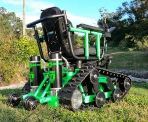 A green and black all-terrain wheelchair with tank-like tracks and large suspension coils sits on grass, designed for outdoor mobility over rough surfaces. Trees and sky are visible in the background.