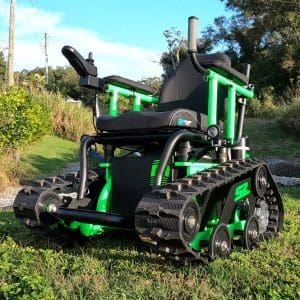 A rugged, all-terrain wheelchair with tank-like tracks and a sturdy black and green frame sits on grass outdoors under a sunny sky, designed for enhanced mobility over rough ground.