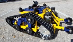 A yellow and blue tracked robotic vehicle with black rubber treads and small wheels at the front, parked on an outdoor asphalt surface. The robot has visible sensors and mechanical components.
