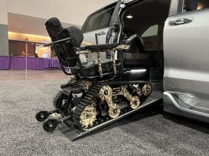 A camouflage-patterned tracked wheelchair is positioned on a ramp extending from the side of a silver minivan at an indoor event. The wheelchair has tank-like treads instead of regular wheels.