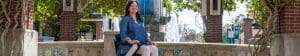 A woman in a blue shirt and white pants sits in a wheelchair by a decorative fountain, surrounded by greenery, brick columns, and tiled artwork in an outdoor plaza.