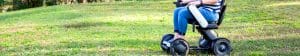 A person wearing blue jeans and a striped shirt sits in a modern motorized wheelchair on a grassy field, with trees visible in the background.