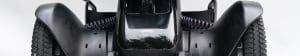 Close-up of the front section of a modern black wheelchair, showing large, textured wheels, smooth curved armrests, and a glossy central panel with the WHILL logo.