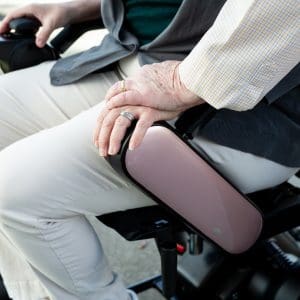 An older adult sits on a mobility scooter, resting a hand on the armrest. The person wears light-colored pants, a green shirt, and a gray vest. The armrest is pink and glossy.