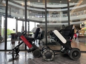 Two modern electric wheelchairs, one red and one white, are parked side by side indoors in front of large glass windows with reflections of a shopping mall interior and people sitting inside.
