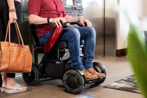 A person in a red shirt and jeans sits in a modern electric wheelchair near an elevator. Another person stands nearby holding a brown handbag. The wheelchair has unique rounded wheels and a sleek design.
