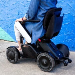 A person wearing a blue shirt and light pants sits in a modern electric wheelchair with large, rugged wheels, positioned on a sidewalk beside a bright blue textured wall.