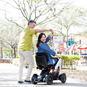 A smiling woman in a blue dress sits in a mobility scooter while a man in a yellow shirt stands beside her, holding her hand as they dance outdoors on a sunny day with trees and red umbrellas in the background.