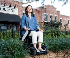 A woman in a blue shirt and white pants smiles while seated on a modern motorized wheelchair outdoors, with a brick building and greenery in the background.