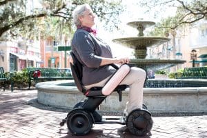 An older woman with gray hair sits in a modern electric wheelchair in front of a fountain, outdoors in a sunny, landscaped urban park.