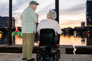 An elderly man and woman enjoy a conversation by a waterfront at sunset. The man stands, smiling, while the woman sits in a mobility scooter. City buildings and colorful lights are visible in the background.