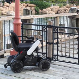 A black electric wheelchair is parked on a wooden boardwalk by a metal gate, overlooking a body of water with rocks and greenery in the background.
