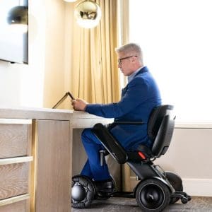 A man in a blue suit sits in a modern electric wheelchair at a desk, using a tablet in a bright room with beige curtains and a spherical hanging light.