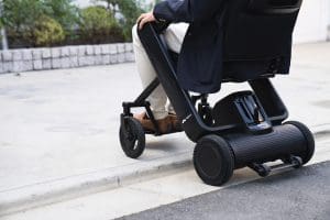 A person in a suit uses a modern motorized wheelchair to navigate up a curb outdoors. The wheelchair has large rear wheels and a sleek, compact design.