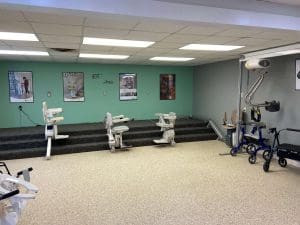 A physical therapy room with exercise equipment, including stair lifts, parallel bars, and walkers. There are three posters on a green wall and the floor is carpeted with a section of black steps.
