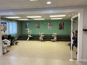 A room with a green accent wall displays three stair lifts on a platform. Posters hang on the wall above the lifts. There are mobility aids, such as wheelchairs and walkers, arranged around the room.