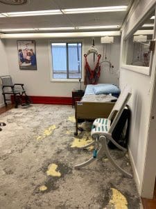 A medical examination room with a hospital bed, medical lift, pillows, a folded shower chair, an office chair, a window, and a wall poster; the floor has a worn, patterned carpet.