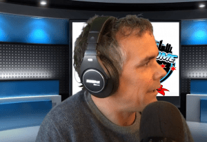 A man wearing large headphones speaks into a microphone in a modern studio with blue lighting and a digital screen displaying a colorful graphic behind him.