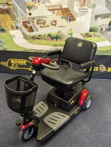 A black and red mobility scooter with a front basket is displayed indoors on a carpet. A colorful accessibility-themed banner is in the background, showing home modifications and labeled features.