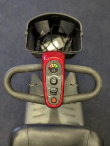 Close-up view of the control panel of a mobility scooter, showing buttons, indicator lights, and a steering handle. A black basket with items inside is attached to the front. The background is out of focus.