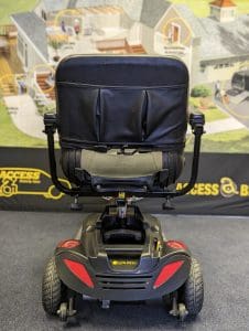 A black mobility scooter viewed from the back, with a cushioned seat and red reflectors, positioned indoors on a carpeted floor. In the background, theres a blurred accessibility-themed display.