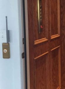 A wooden elevator door is partially open, revealing an elevator control panel with buttons and a red emergency switch inside. A brass call button panel is mounted on the wall next to the door.