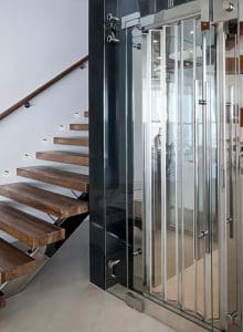 A modern interior featuring floating wooden stairs with metal supports next to a sleek glass elevator with metal accents and a reflective surface.
