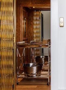 A home elevator with wooden paneling and a gold accordion gate is open, revealing a metal rolling cart inside with two large silver bowls placed on its lower shelf.