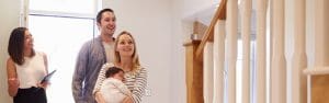 A smiling couple with a baby in their arms stands inside a bright home, looking up the staircase, while a woman holding a tablet, possibly a real estate agent, stands nearby and gestures.