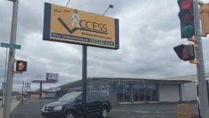 A yellow and black ACCESS business sign stands next to a street intersection with a traffic light and walk signals. A black van is parked in front of a modern, glass-fronted building under a cloudy sky.