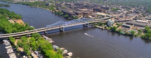 Aerial view of a city with a blue truss bridge crossing a wide river, boats docked along the shores, and a boat leaving a wake; buildings and roads surround the bridge, highlighting La Crosse accessibility with visible wheelchair ramps in Wisconsin.