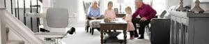 Two older adults and two children sit together in a bright living room, playing at a wooden coffee table. A stairlift is visible on the left side of the image near a staircase.