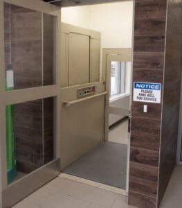 A V-1504 Vertical Platform Lift by Savaria sits beside a wood-paneled wall with a "NOTICE: Please ring bell for service" sign in a corridor with tiled floors, providing a smooth and reliable platform lift experience.