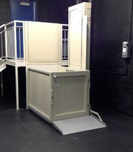 A Savaria V-1504 Vertical Platform Lift with a smooth ride platform and ramp is installed indoors next to stairs and a metal railing, surrounded by dark walls and flooring.