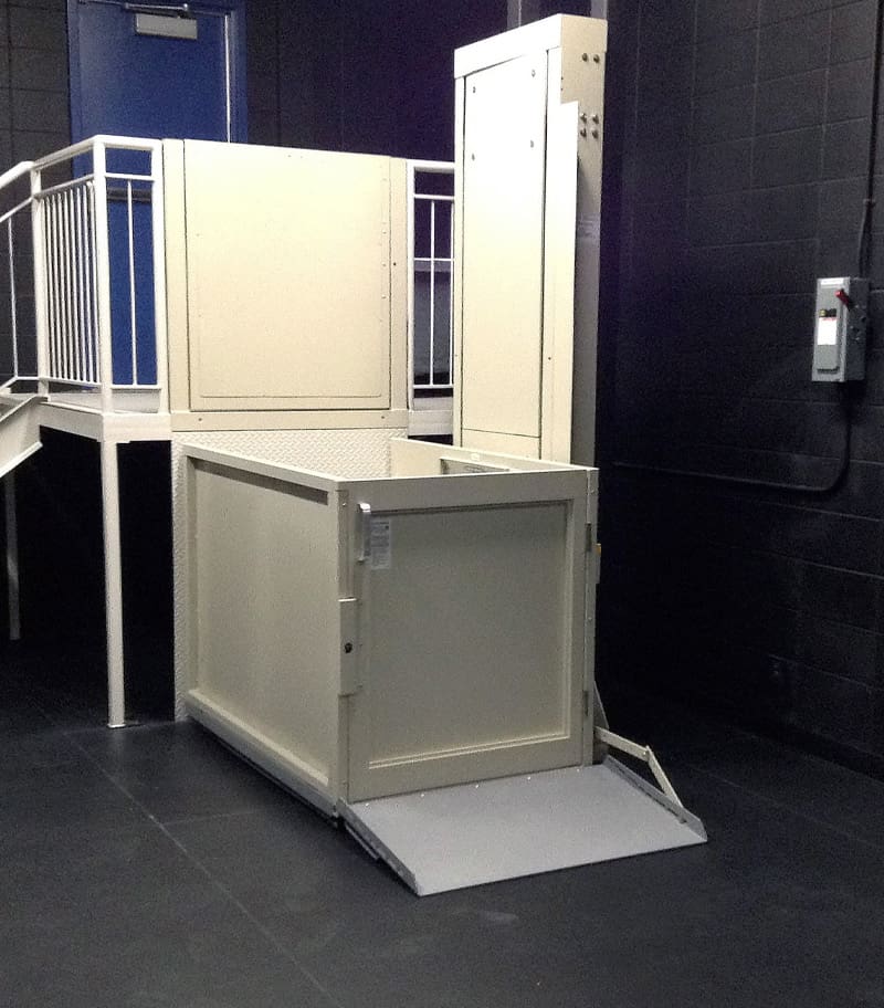 A Savaria V-1504 Vertical Platform Lift with a smooth ride platform and ramp is installed indoors next to stairs and a metal railing, surrounded by dark walls and flooring. A Savaria V-1504 Vertical Platform Lift with a smooth ride platform and ramp is installed indoors next to stairs and a metal railing, surrounded by dark walls and flooring.